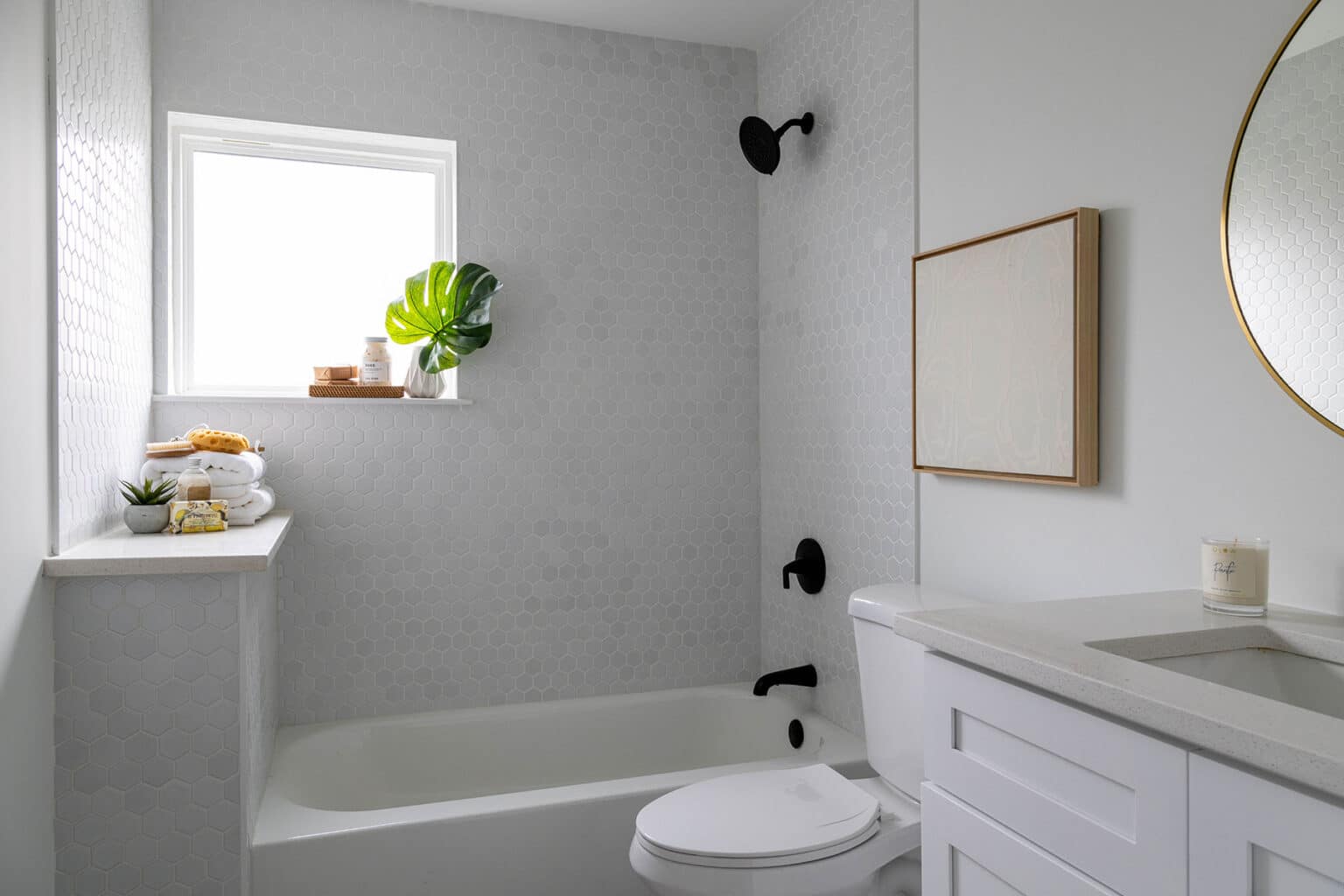 Minimalist bathroom featuring a white bathtub and shower combination with light textured tile walls and matte black fixtures. A small window above the tub lets in natural light and holds a wooden tray with a plant and toiletries. The tub area includes a ledge with folded towels, a candle, and a small potted succulent. To the right, a white vanity with a light stone countertop is topped with a candle, and a round mirror with a thin brass frame hangs above it. A simple square wall art piece complements the clean, modern aesthetic of the space.