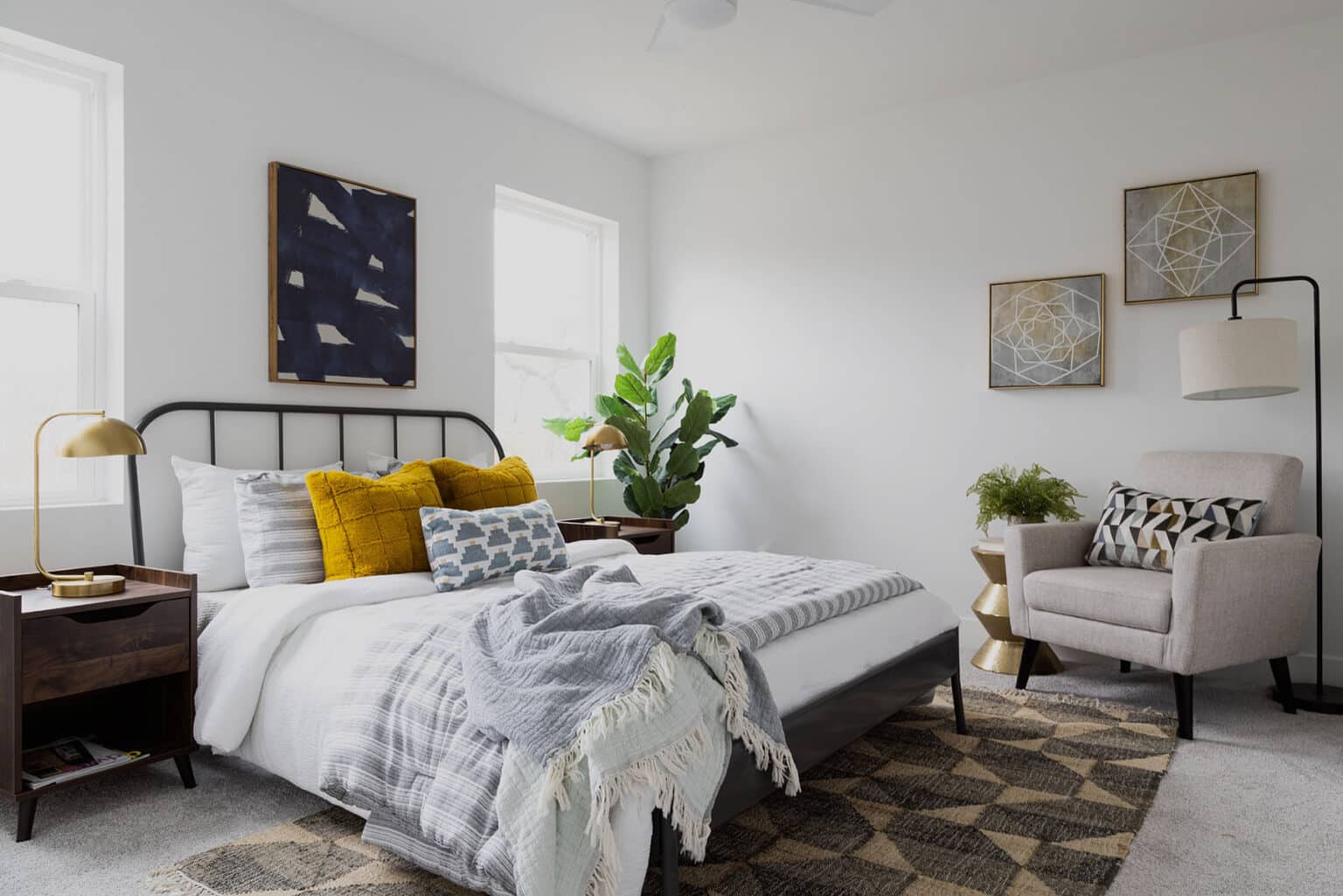 Modern and cozy bedroom with a minimalist design. The space features a low-profile bed with a black metal frame, white and gray bedding, and accent pillows in mustard yellow and geometric patterns. Flanking the bed are two dark wood nightstands with gold lamps. A large leafy plant adds greenery near the corner window. On the right, a light beige armchair with a patterned pillow sits beside a sculptural gold side table and a black floor lamp. Abstract artwork decorates the white walls, while a textured area rug adds warmth to the carpeted floor. Natural light fills the room through two large windows.