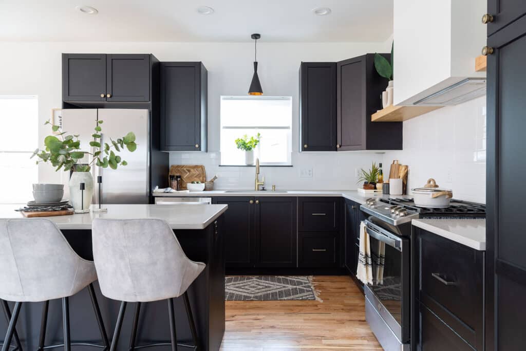 Modern kitchen with a bold contrast of black cabinetry and white countertops. The space features a U-shaped layout with stainless steel appliances, including a gas range and refrigerator. A central window above the sink allows natural light to brighten the room, complemented by a single black pendant light overhead. The island includes two gray upholstered barstools and is decorated with a vase of greenery and stacked dishes. White backsplash tiles, warm wood flooring, and brass fixtures add warmth and elegance to the sleek, contemporary design.
