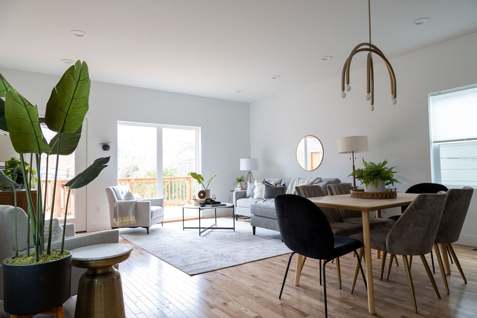 Open-concept living and dining area with a bright, airy atmosphere and contemporary decor. The living space features a light gray sectional sofa with accent pillows, two armchairs, and a sleek coffee table on a large area rug. A round mirror and floor lamp add style and functionality. Adjacent to the seating area is a light wood dining table surrounded by a mix of black and gray upholstered chairs. A sculptural modern chandelier hangs above the table. Large windows and sliding glass doors let in natural light, while tall potted plants bring in a fresh, organic touch. Warm wood flooring ties the space together.