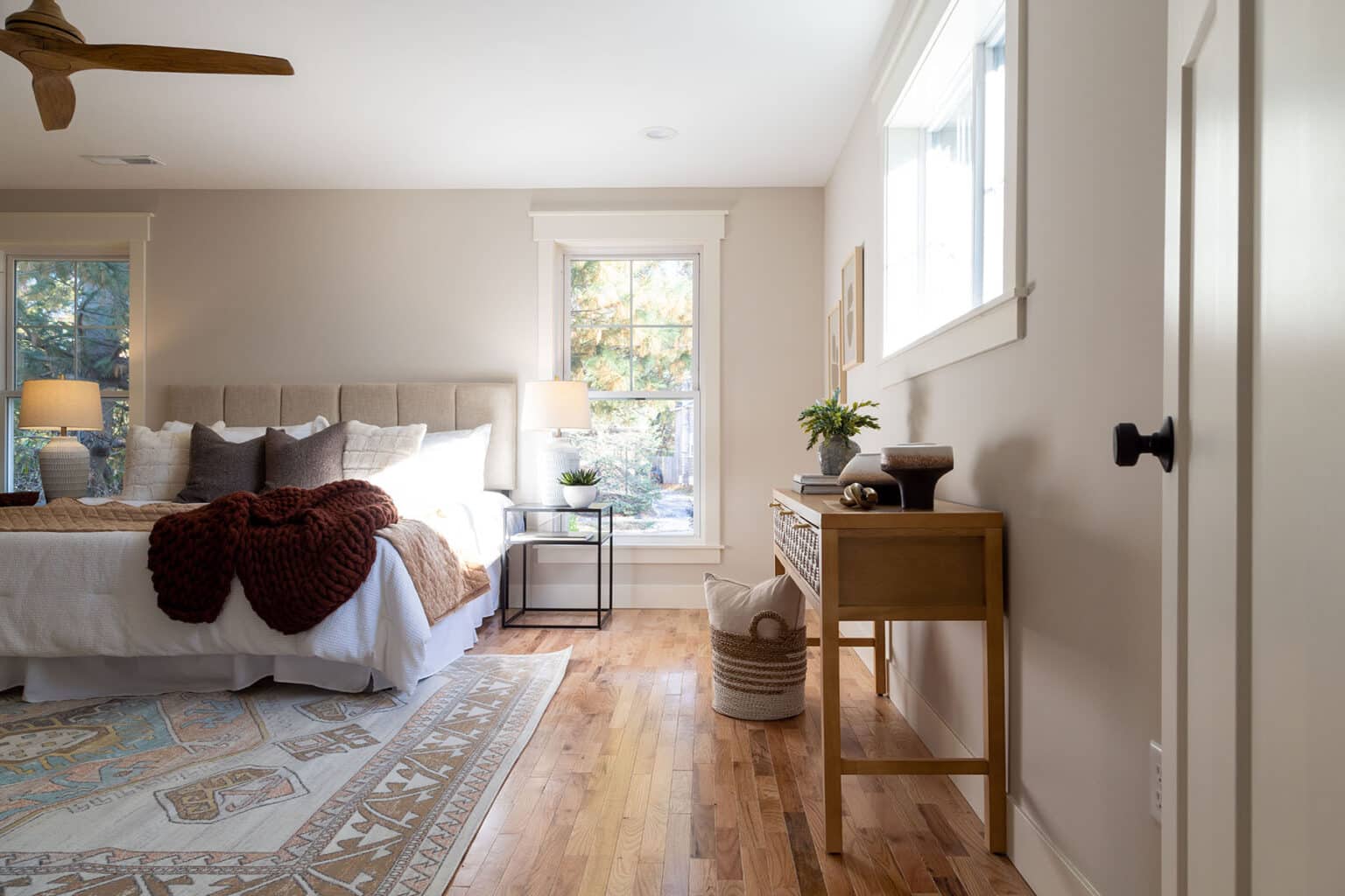 Bright and serene bedroom featuring a neutral color palette and natural wood accents. A tufted upholstered bed is dressed in crisp white bedding with layered throw pillows and a deep burgundy knit blanket. Two modern black-framed side tables with matching lamps flank the bed. A large patterned area rug adds softness over the warm hardwood floor. Along the opposite wall, a light wood console table is styled with decorative objects, books, and a small potted plant. A woven basket with blankets sits on the floor below. Multiple windows fill the room with natural light, enhancing the cozy, inviting atmosphere.