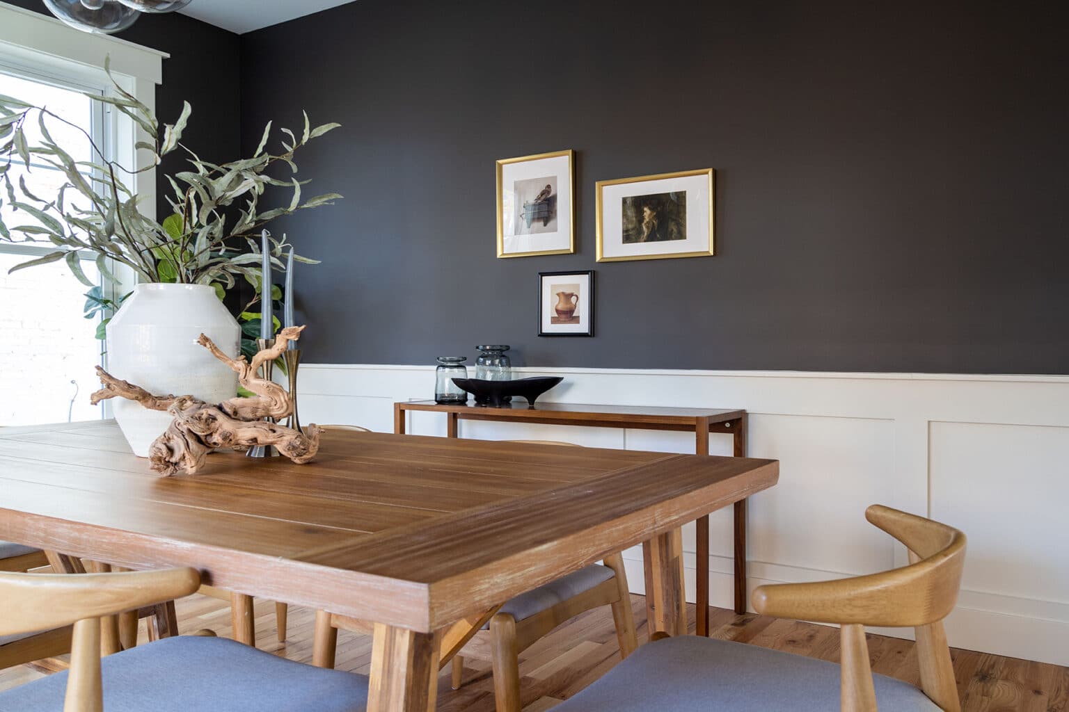 Modern dining room with a minimalist yet inviting aesthetic. The space features a natural wood dining table surrounded by matching wooden chairs with light gray cushions. A large white ceramic vase with greenery and a sculptural wood centerpiece decorates the table. The walls are painted a dramatic charcoal color above crisp white wainscoting. On the back wall, three framed art prints in gold frames add a touch of elegance. A narrow wood sideboard holds simple black serveware and glass jars, contributing to the clean and cohesive design. Natural light filters in from a nearby window, enhancing the warmth of the wood flooring.