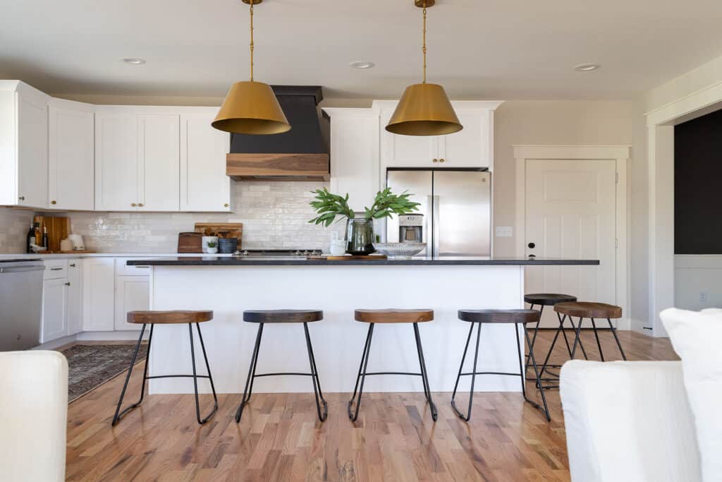 Bright and open kitchen with a large island featuring a dark countertop and seating for six with wooden barstools and black metal legs. The kitchen is styled with white shaker cabinets, stainless steel appliances, and a striking black and wood range hood above the stove. Two oversized brass pendant lights hang above the island, adding a touch of warmth and elegance. Light wood flooring and neutral walls enhance the airy, welcoming feel of the space, which opens into a larger living area. A pair of glass vases with greenery sit atop the island for a fresh, decorative touch.