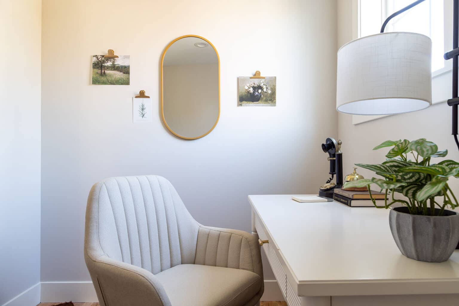 Chic and compact home office nook with a clean, minimal aesthetic. A white desk is paired with a plush, vertically channeled upholstered chair in a soft beige tone. On the desk sits a modern black lamp, a small stack of books, and a potted plant for a touch of greenery. Above the desk, a gold-framed oval mirror is flanked by small clipped photographs and art prints. A window on the right lets in natural light, enhancing the room’s airy and inviting feel.
