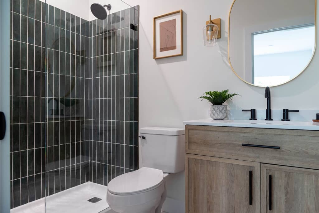 Modern bathroom with a sleek, stylish design. The walk-in shower features dark vertical subway tiles and a frameless glass door with a matte black showerhead. Next to the shower is a white toilet, and beside it, a light wood vanity with black hardware and a white countertop. The vanity includes a black faucet and a small potted plant for a natural touch. Above the sink is a round mirror with a thin gold frame, paired with a wall-mounted sconce. Minimalist artwork and natural light enhance the clean, contemporary feel of the space.