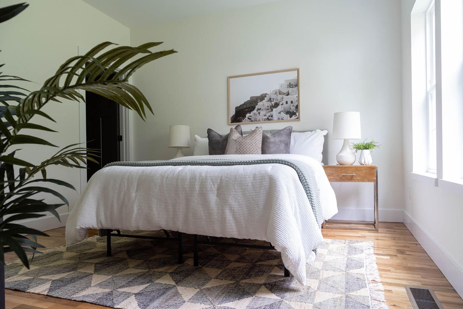Bright and serene bedroom with a minimalist design. The space features a bed with white bedding, textured throw pillows in neutral tones, and a gray accent blanket. A wooden nightstand with a white lamp and greenery sits on each side of the bed. Above the headboard hangs a framed black-and-white photograph of a cliffside village. The room is grounded by a geometric area rug on warm hardwood floors, and natural light pours in from large windows on the right. A tall green plant in the foreground adds a touch of life and contrast to the soft, calming palette.