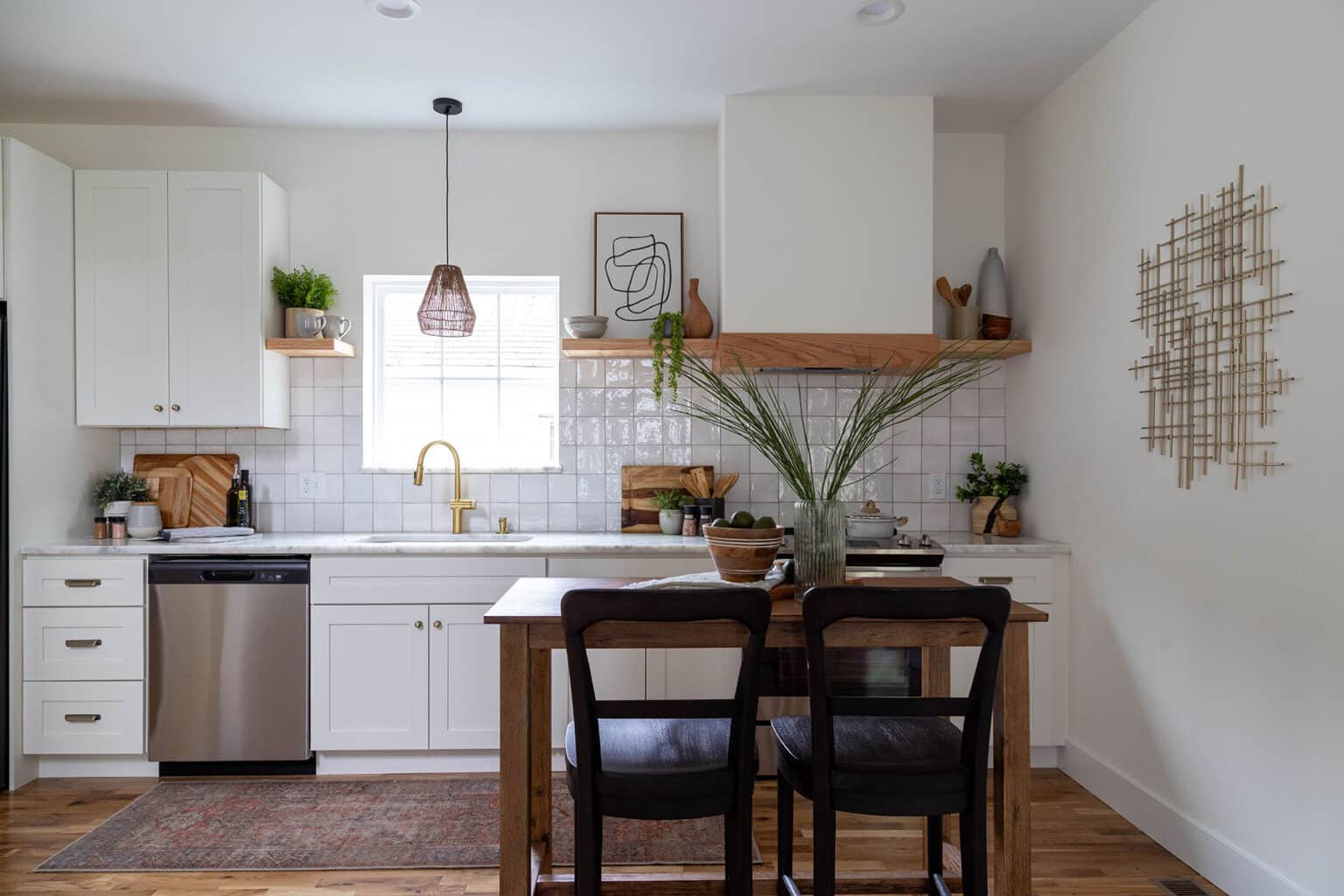 Charming and cozy kitchen with a mix of modern and rustic elements. White cabinetry and square tile backsplash are complemented by warm wood accents, including floating shelves and a central dining table. A brass faucet sits beneath a window that brings in natural light, with a woven pendant light hanging above. The wooden dining table is set with a glass vase holding tall greenery and paired with dark wooden chairs. Open shelving displays minimal decor, plants, and pottery, adding character and warmth to the space. A stainless steel dishwasher and subtle wall art complete the inviting kitchen design.