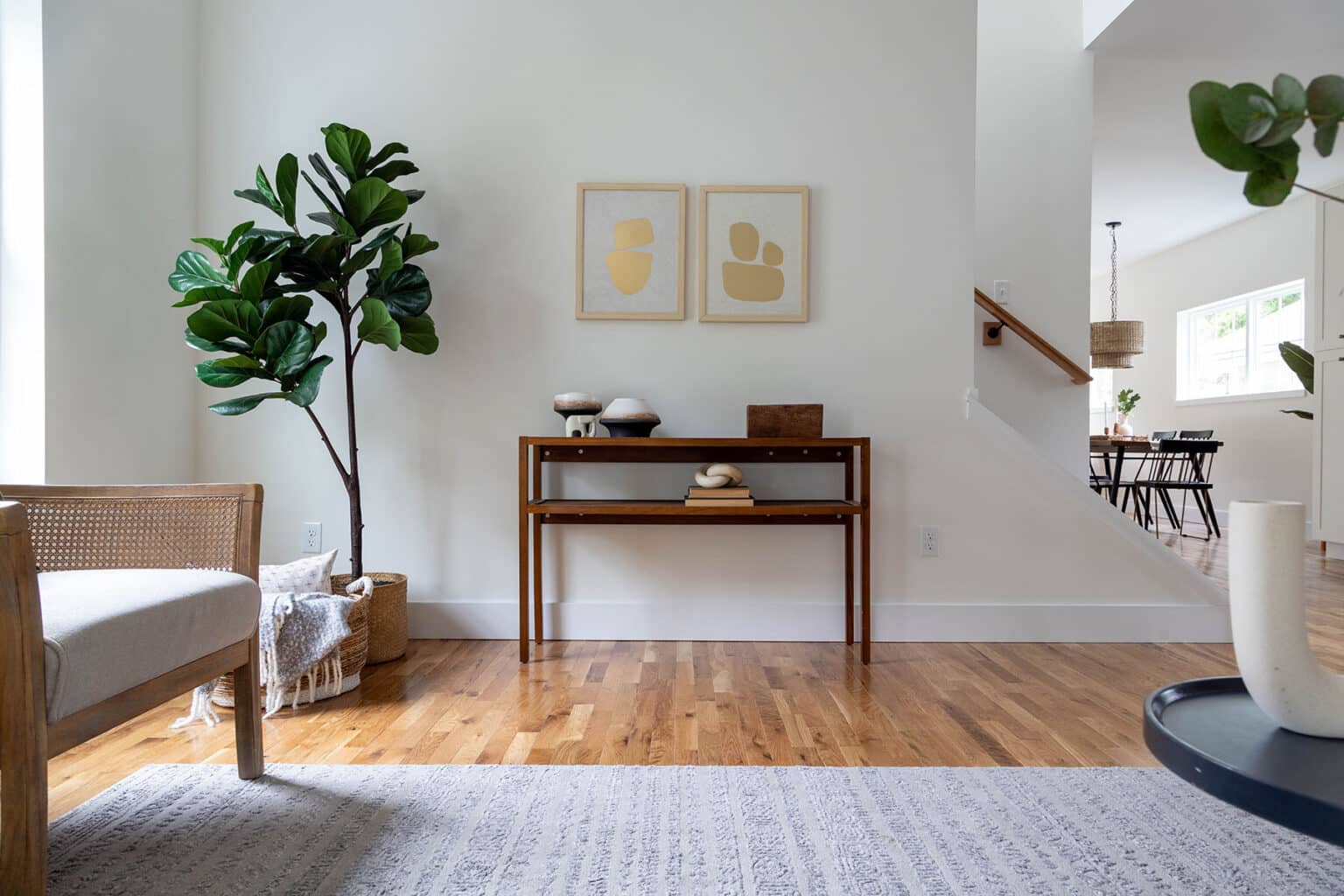 Modern and airy living space with warm wood flooring and a minimalist aesthetic. A tall fiddle-leaf fig tree in a woven basket adds a natural touch in the corner, next to a light wood chair with a cane back and neutral cushion. A wooden console table along the wall is styled with decorative objects, including ceramic vessels, stacked books, and a sculptural piece. Above the table, two abstract art prints in muted gold tones add visual interest. Soft natural light filters into the space, which connects to an open dining area visible in the background. A textured area rug anchors the cozy seating area in the foreground.