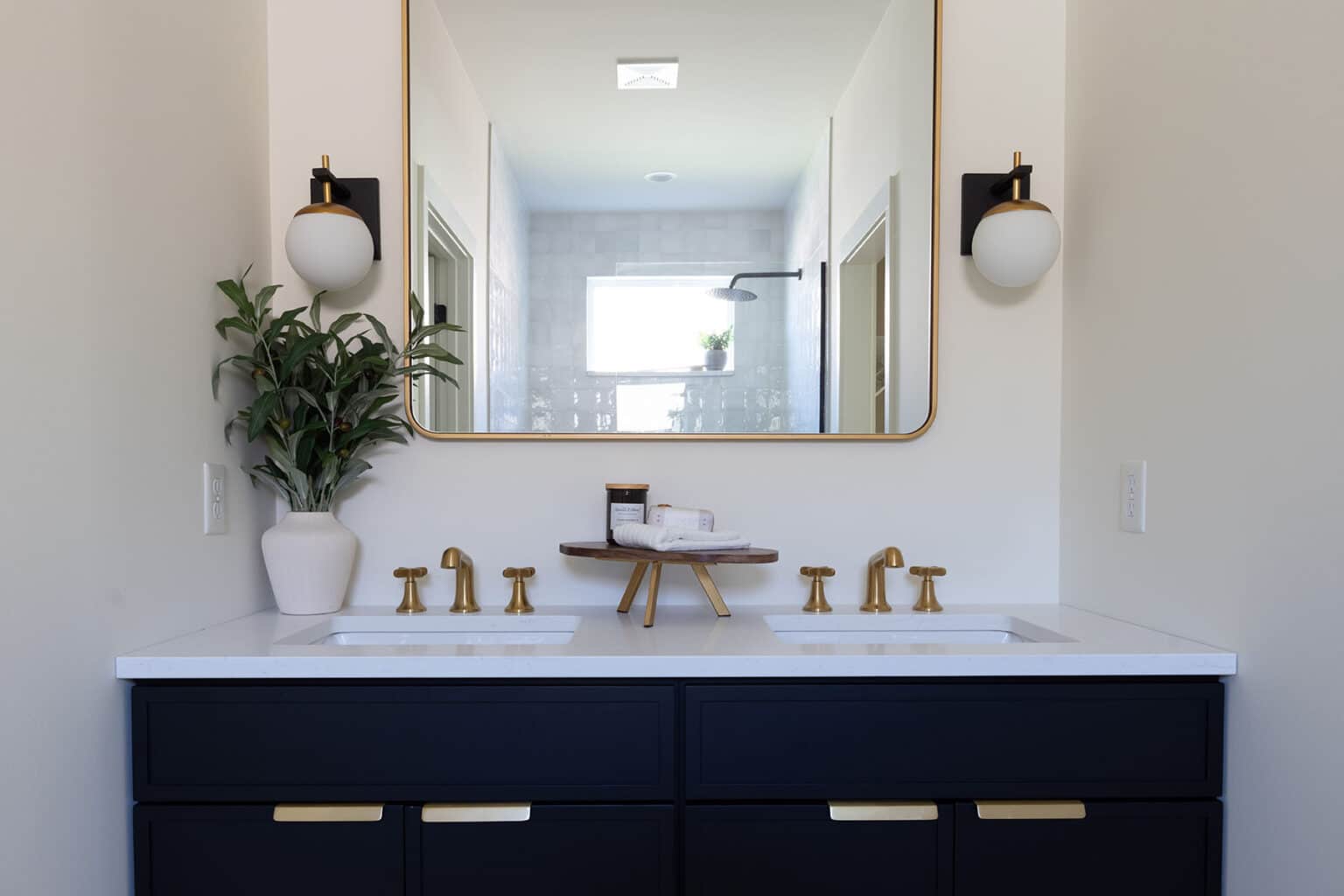 Elegant double vanity bathroom featuring a sleek black cabinet with gold hardware and white countertops. Each sink is paired with a matching gold faucet. Above the vanity hangs a large rectangular mirror with a thin gold frame, flanked by two modern globe sconces. A potted plant adds greenery to the left side of the counter, while a small wooden tray holds toiletries at the center. The mirror reflects a glimpse of the walk-in shower with a window letting in natural light, enhancing the bathroom’s bright and polished aesthetic.