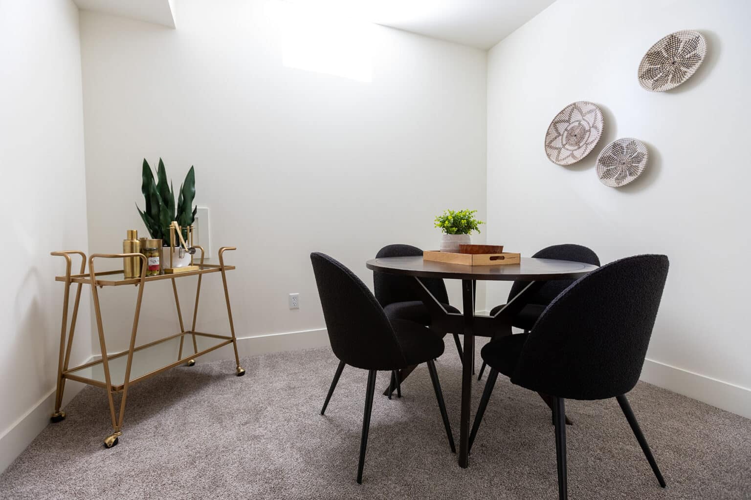Stylish and compact dining or meeting area with a round black table surrounded by five black upholstered chairs. The room features soft gray carpeting and white walls, creating a clean and modern aesthetic. A trio of decorative woven baskets is arranged as wall art. To the left, a gold-framed bar cart with glass shelves holds drinks, decor, and a potted snake plant. A small wooden tray with a green plant sits at the center of the table, adding a touch of warmth and freshness to the minimalist space.