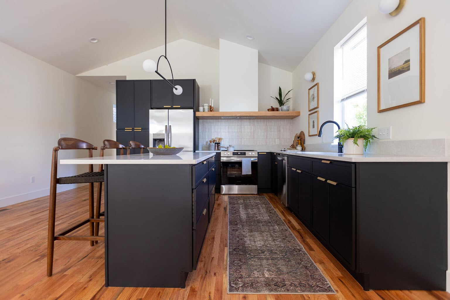 Contemporary kitchen with a vaulted ceiling and a sleek, modern design. The space features matte black lower cabinets with gold hardware, white countertops, and a white tile backsplash behind a stainless steel range. A wooden open shelf above the stove displays minimal decor. A long patterned runner rug lies on the warm wood floor, and a modern black pendant light hangs above the island with seating for two. Wall-mounted artwork and potted plants add a touch of warmth and character. Natural light streams in from a tall window on the right.