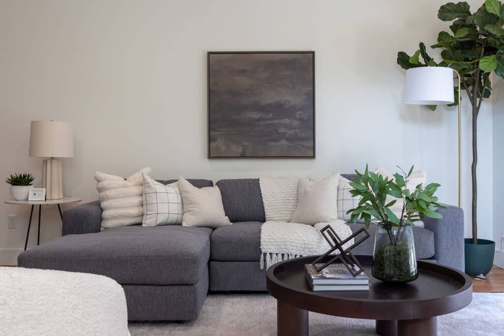 Modern living room with a cozy, neutral palette. A dark gray sectional sofa is adorned with textured cream and gray throw pillows and a soft knit blanket. A round dark wood coffee table sits in front of the sofa, styled with a vase of greenery, a few stacked books, and a decorative metal sculpture. On the left, a small round side table holds a white lamp and a potted plant. A large abstract dark-toned artwork hangs above the sofa, and a tall fiddle-leaf fig tree and floor lamp complete the right corner, adding height and warmth to the space.