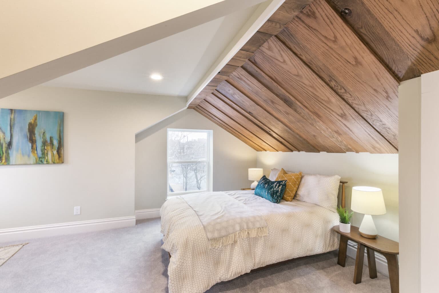 A carpeted room with a slanted, wooden ceiling includes a well-made bed, side table, and wall art.