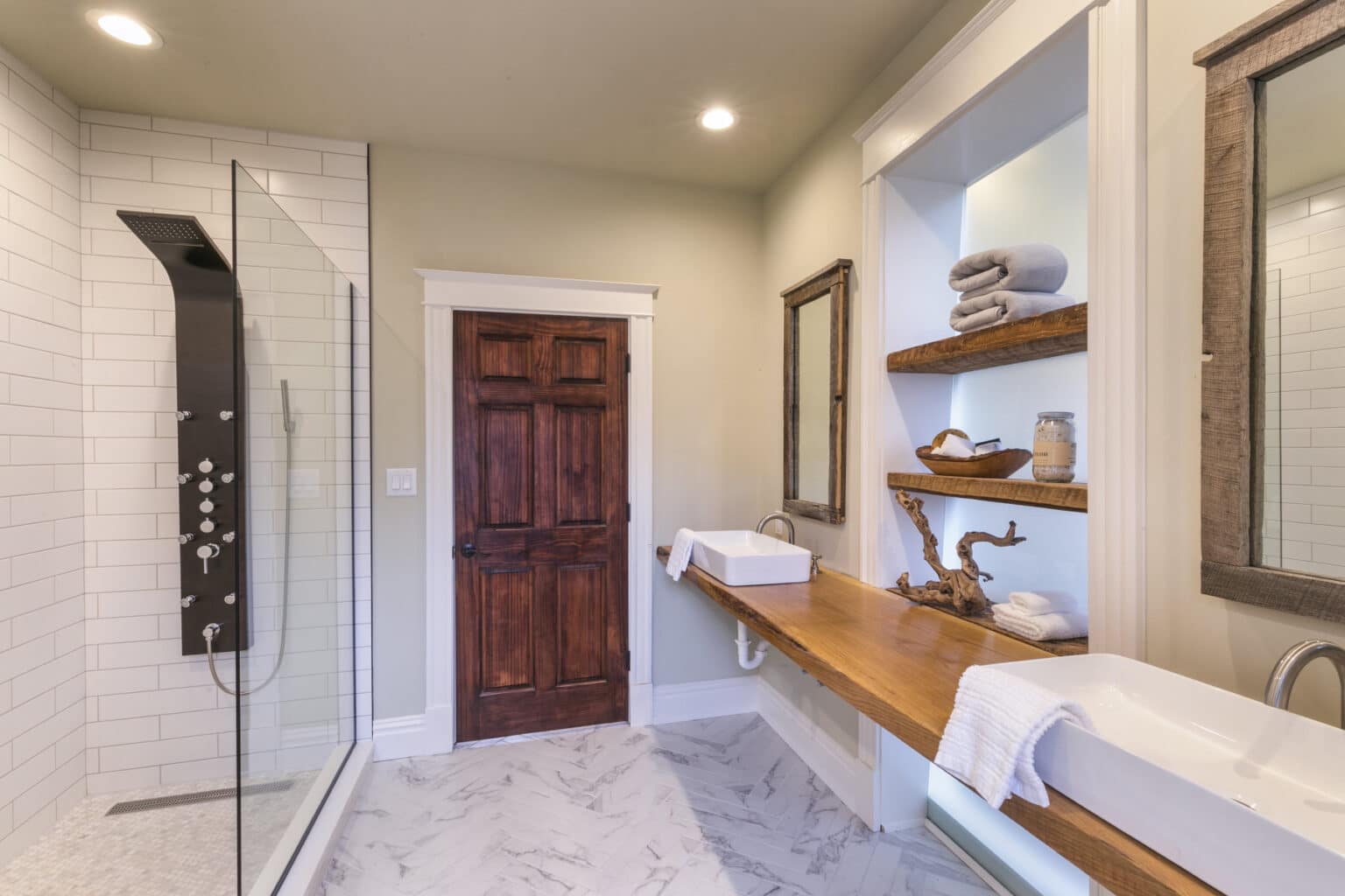 A modern bathroom with tiled shower, wooden vanity and two sinks is tastefully decorated.