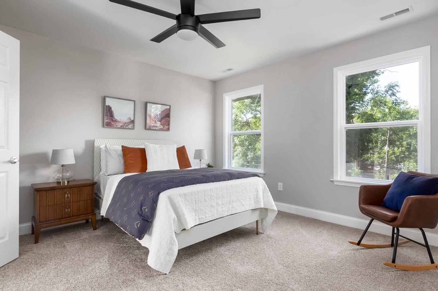 A carpeted bedroom with two large windows, well-made bed, and sitting area.