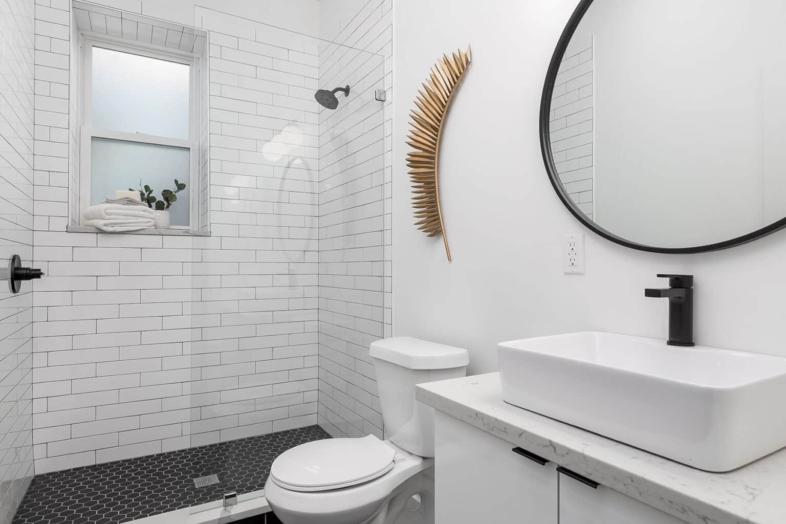 Modern minimalist bathroom featuring a walk-in shower with white subway tile walls and dark hexagon floor tiles. A frosted window above the shower holds folded white towels and a small potted plant. The vanity has a white rectangular vessel sink, black faucet, and white countertop with black hardware. A round black-framed mirror hangs above the sink, and a decorative gold palm leaf wall sculpture adds a touch of warmth to the white wall.