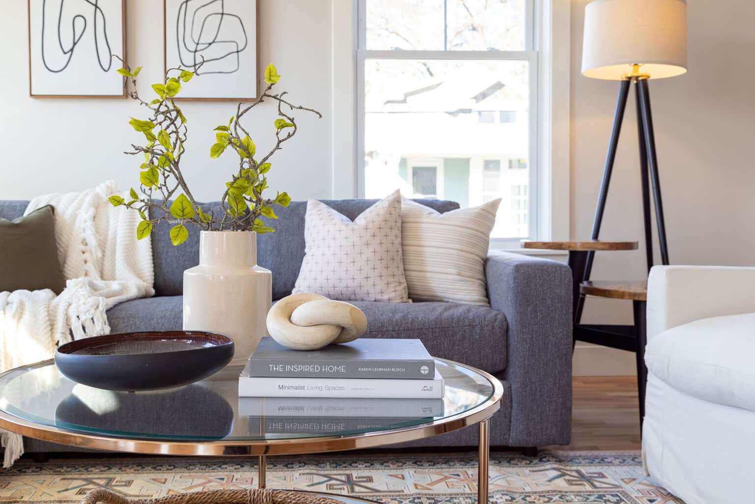Stylish and inviting living room featuring a dark gray sofa adorned with neutral throw pillows and a cozy knit blanket. In front of the sofa is a round glass coffee table with a wooden base, topped with decorative books, a modern ceramic sculpture, a dark bowl, and a beige vase holding branches with green leaves. A patterned area rug adds texture beneath the table. To the right, a white armchair and a tripod floor lamp with a white shade complete the space. Large windows bring in natural light, highlighting the warm and modern decor.