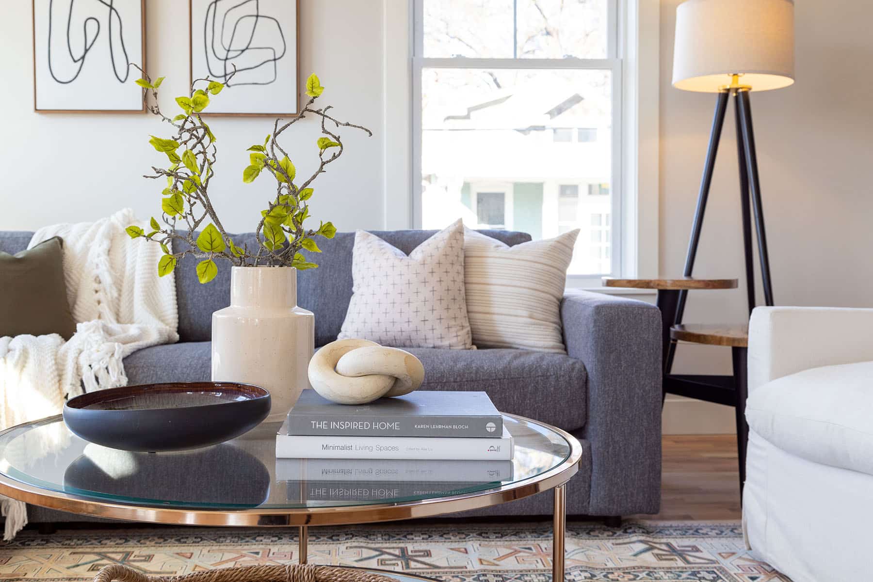 Stylish and inviting living room featuring a dark gray sofa adorned with neutral throw pillows and a cozy knit blanket. In front of the sofa is a round glass coffee table with a wooden base, topped with decorative books, a modern ceramic sculpture, a dark bowl, and a beige vase holding branches with green leaves. A patterned area rug adds texture beneath the table. To the right, a white armchair and a tripod floor lamp with a white shade complete the space. Large windows bring in natural light, highlighting the warm and modern decor.