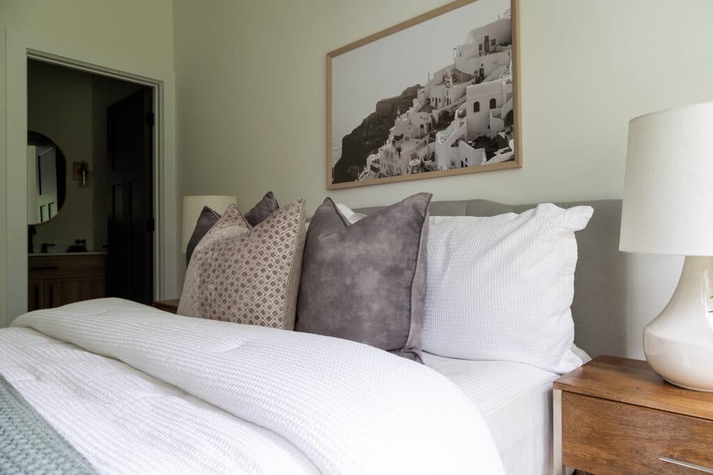 Cozy, modern bedroom featuring a neatly made bed with white bedding and a mix of textured and patterned throw pillows in neutral tones. A wooden nightstand with a ceramic lamp sits on either side of the bed. Above the bed hangs a framed photograph of white cliffside buildings in Santorini, Greece. The room has soft, muted wall colors and a doorway leading to a bathroom with a round mirror visible in the background.