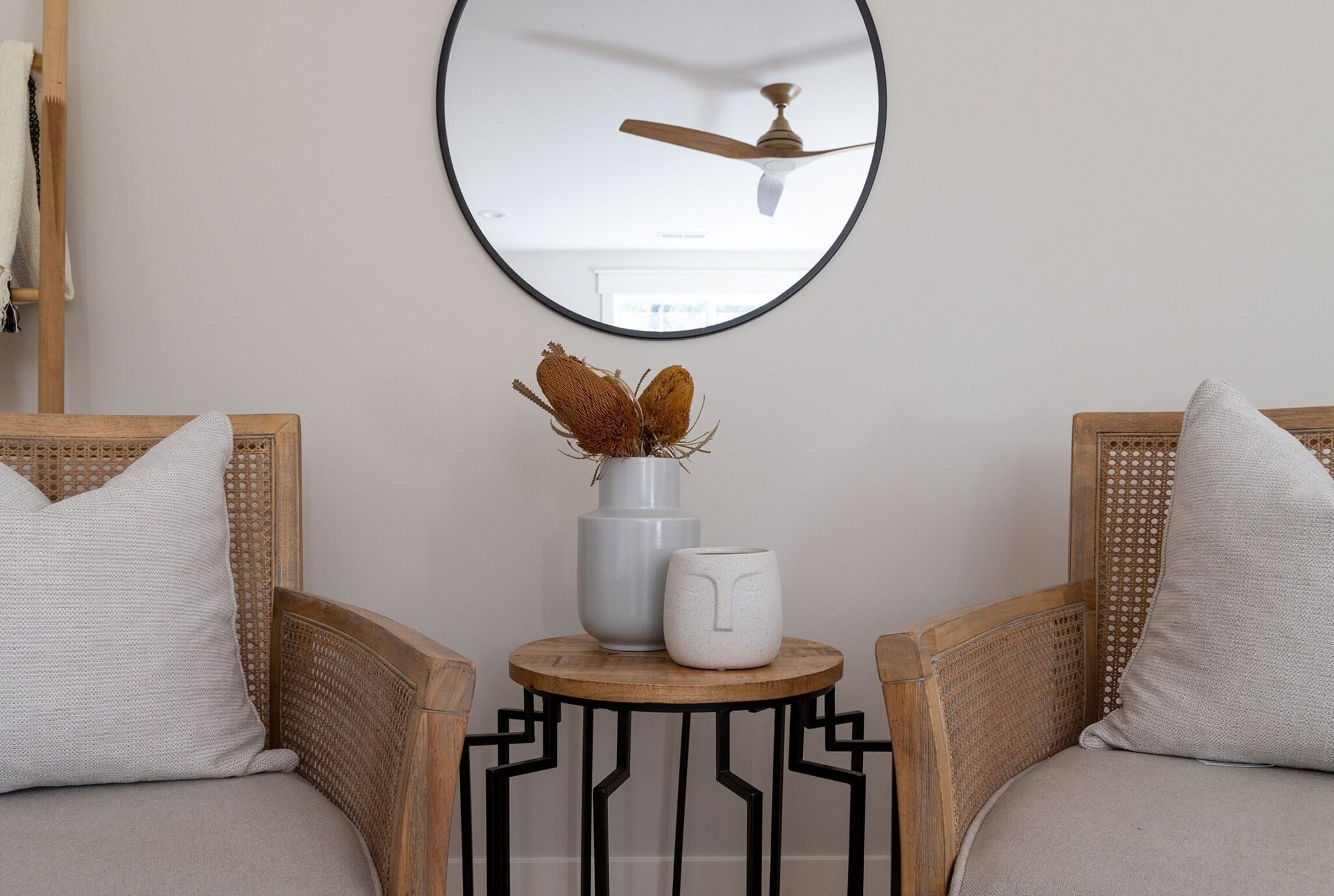 Neutral-toned seating area featuring two wooden armchairs with woven cane backs and light gray cushions, each accented with a soft throw pillow. Between the chairs is a small round wooden side table with black metal legs, topped with minimalist ceramic vases and dried floral stems. A round black-framed mirror above the table reflects a ceiling fan and adds depth to the light beige wall. A wooden blanket ladder with folded textiles leans against the wall to the left, enhancing the cozy, modern aesthetic.