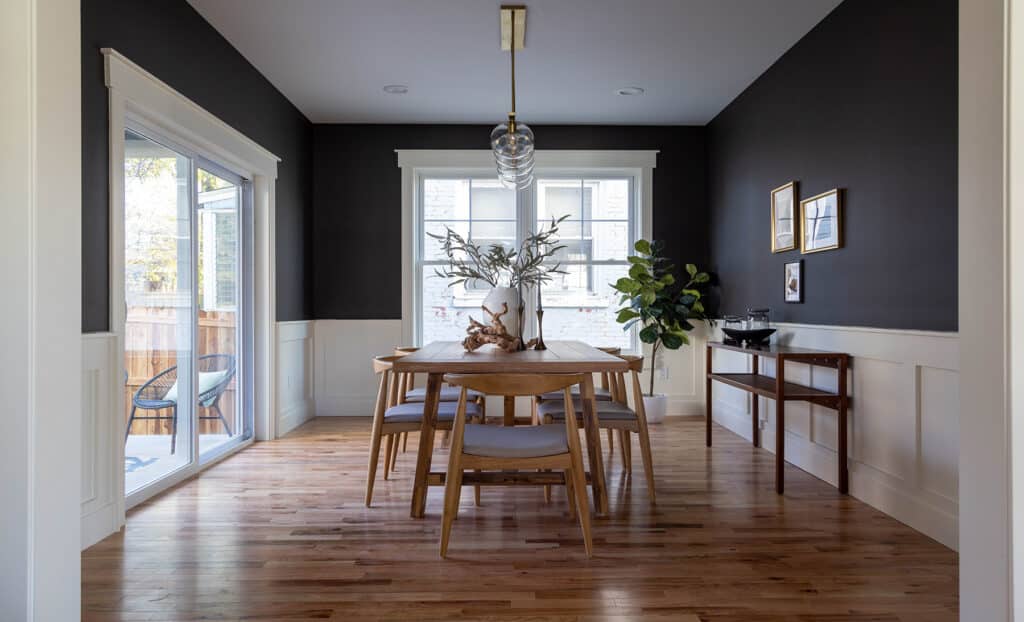 Elegant dining room featuring dark charcoal upper walls with crisp white wainscoting below. A light wood dining table is surrounded by six matching mid-century modern chairs with light gray cushions. Centered above the table hangs a modern glass pendant light. The room is brightened by large windows and a sliding glass door that opens to a deck. A sideboard with minimal decor and framed artwork adds balance to the space, while a potted fiddle-leaf fig tree in the corner brings a natural touch. Warm hardwood floors complete the sophisticated yet inviting atmosphere.