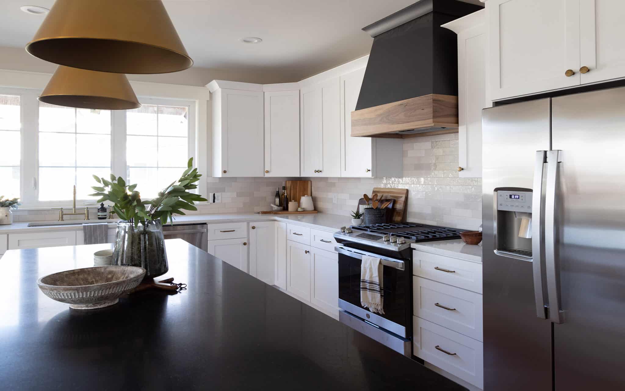 Modern farmhouse kitchen with a clean and sophisticated design. The space features white shaker cabinets with brass hardware, a large stainless steel refrigerator, and a matching gas range with a striking black and wood range hood above. A dark quartz island countertop contrasts with the white cabinetry, decorated with glass vases, a decorative bowl, and greenery. Two large tan pendant lights hang above the island. White subway tile backsplash and ample natural light from large windows above the farmhouse sink complete the airy and inviting atmosphere.