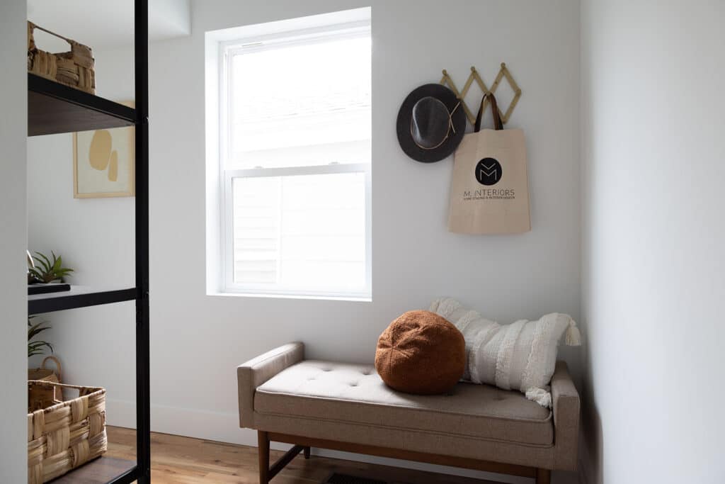 Bright and cozy entryway or nook featuring a light gray tufted bench with wooden legs, accented by a round rust-colored pillow and a textured white cushion. A window above the bench lets in natural light, creating an airy atmosphere. On the wall above the bench, a folding accordion-style wall hook holds a dark felt hat and a canvas tote bag labeled “M. Interiors.” To the left, black metal shelves with woven baskets and plants add a touch of warmth and functionality to the space.