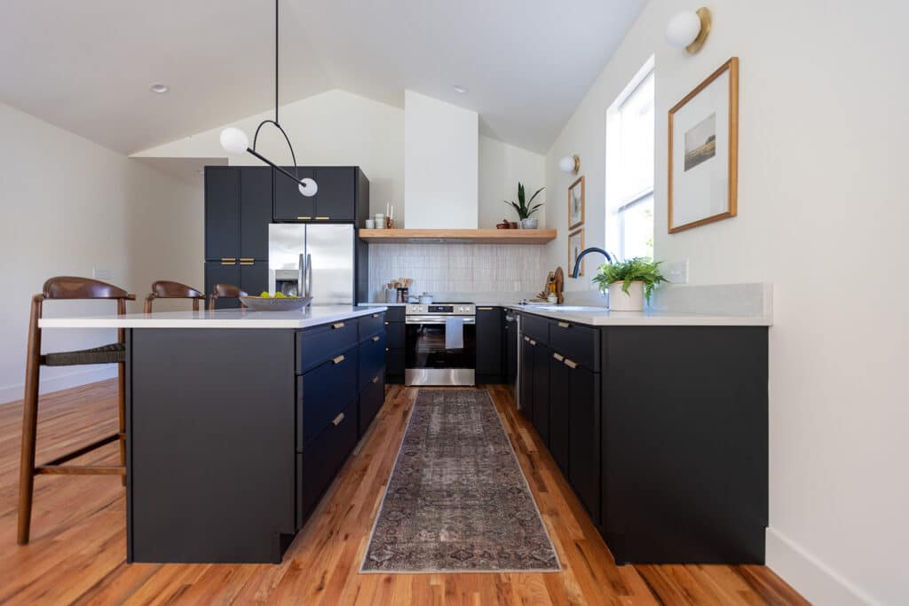 Contemporary kitchen with a vaulted ceiling and a sleek, modern design. The space features matte black lower cabinets with gold hardware, white countertops, and a white tile backsplash behind a stainless steel range. A wooden open shelf above the stove displays minimal decor. A long patterned runner rug lies on the warm wood floor, and a modern black pendant light hangs above the island with seating for two. Wall-mounted artwork and potted plants add a touch of warmth and character. Natural light streams in from a tall window on the right.