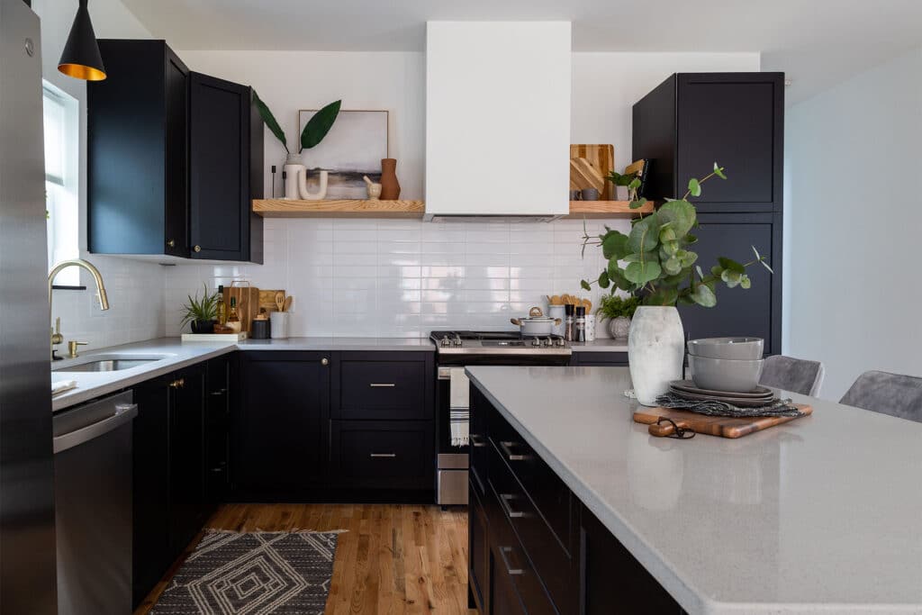 Contemporary kitchen with a bold, elegant design featuring matte black cabinets, brass hardware, and light quartz countertops. A large island with seating offers space for dining and prep, topped with a vase of fresh greenery and stacked dishware. The backsplash is composed of sleek white subway tiles, and open wood shelving above the stove displays decorative items and kitchen essentials. Natural wood floors add warmth, while gold fixtures and black pendant lighting enhance the upscale, modern aesthetic.