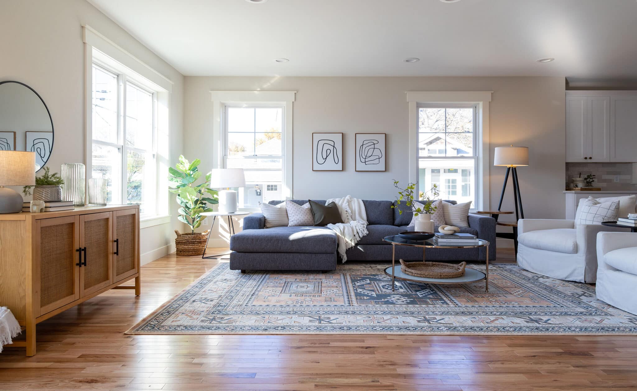 Spacious and inviting living room filled with natural light from multiple large windows. A dark gray sectional sofa is complemented by neutral-toned throw pillows and a cozy blanket, while two white swivel chairs sit opposite. A large patterned area rug anchors the seating area, with a round glass coffee table at the center topped with decorative books and a vase of greenery. The room features warm hardwood floors, modern abstract artwork, and layered lighting from table and floor lamps. A wooden sideboard with a round mirror and styled decor adds warmth and texture to the left side of the space.
