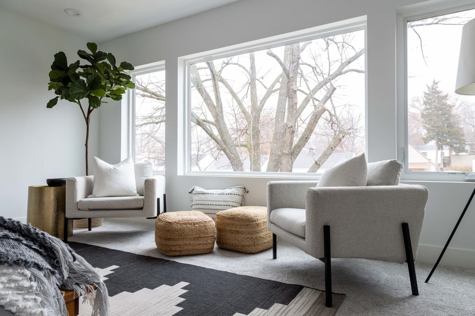Bright and cozy sitting area with large windows showcasing a view of bare winter trees. The space features two modern light-gray armchairs with white throw pillows, flanking two woven rattan poufs that serve as ottomans or casual seating. A black-and-white geometric rug adds contrast over the light carpeted floor. A tall leafy plant in the corner and a sleek floor lamp complete the corner nook, creating a serene and stylish spot for relaxation or conversation.