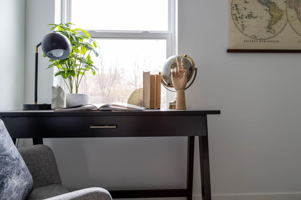 Modern home office corner featuring a sleek black desk positioned in front of a window with a view of bare trees. The desktop is styled with an open book, a black and gold desk lamp, a potted plant, and a globe. A set of vertical books is held by a decorative wooden mannequin hand, adding a creative touch. To the right, part of a vintage-style world map hangs on the wall. A gray upholstered chair sits just in frame, completing the cozy and stylish workspace.