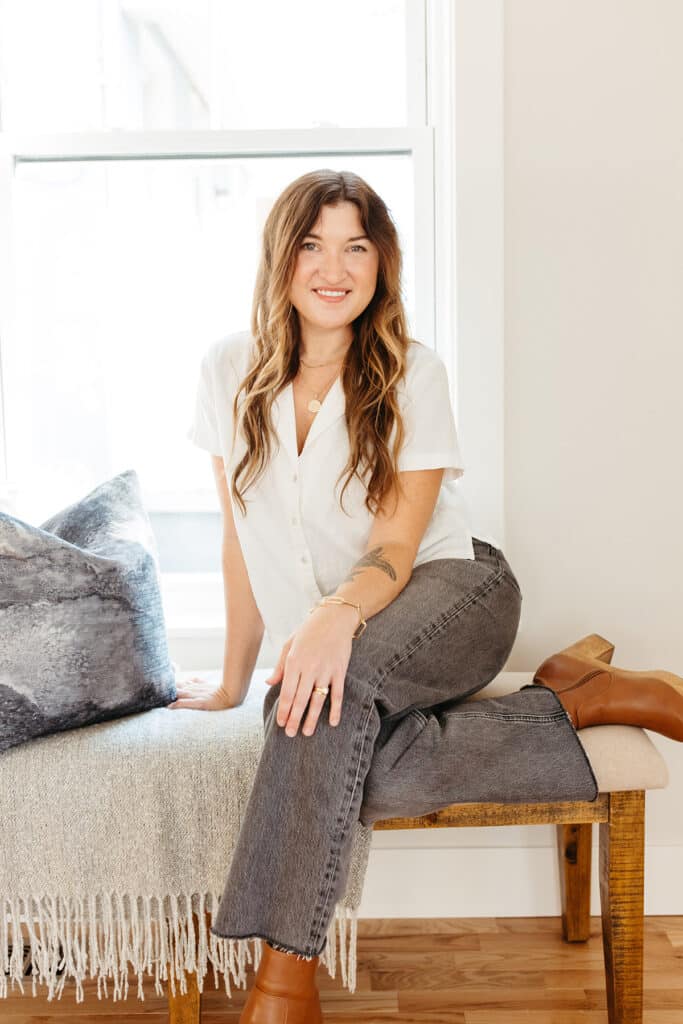 A woman with long wavy brown hair sits on a wooden bench in a bright room with natural light streaming through a large window behind her. She is smiling and casually dressed in a white short-sleeve button-up shirt, gray jeans, and brown leather ankle boots. The bench is topped with a soft gray throw and a decorative gray pillow. The overall setting is warm and inviting with neutral tones and a cozy, modern feel.