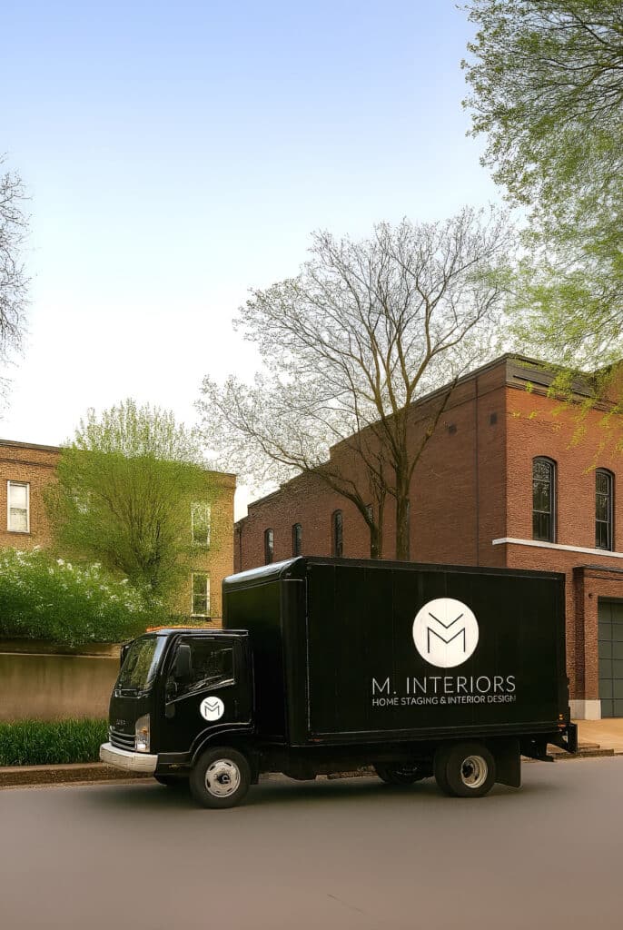 A sleek black box truck with the “M. INTERIORS – Home Staging & Interior Design” logo and text in white is parked on a quiet, tree-lined street. The truck features a large circular logo on the side and a smaller version on the driver’s door. Behind it are historic red-brick and tan-brick buildings, with budding spring trees and greenery under a clear blue sky.